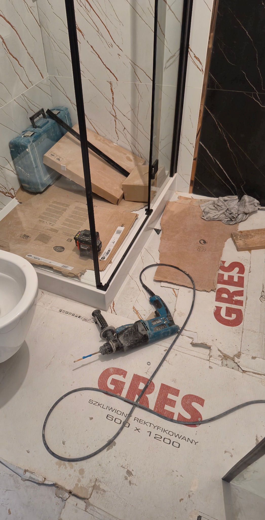 Montaż kabiny prysznicowej: widoczna wiertarka udarowa Makita na podłodze wyłożonej płytkami gresowymi, częściowo zabezpieczonej kartonem. W tle niebieska skrzynka narzędziowa i poziomica laserowa.