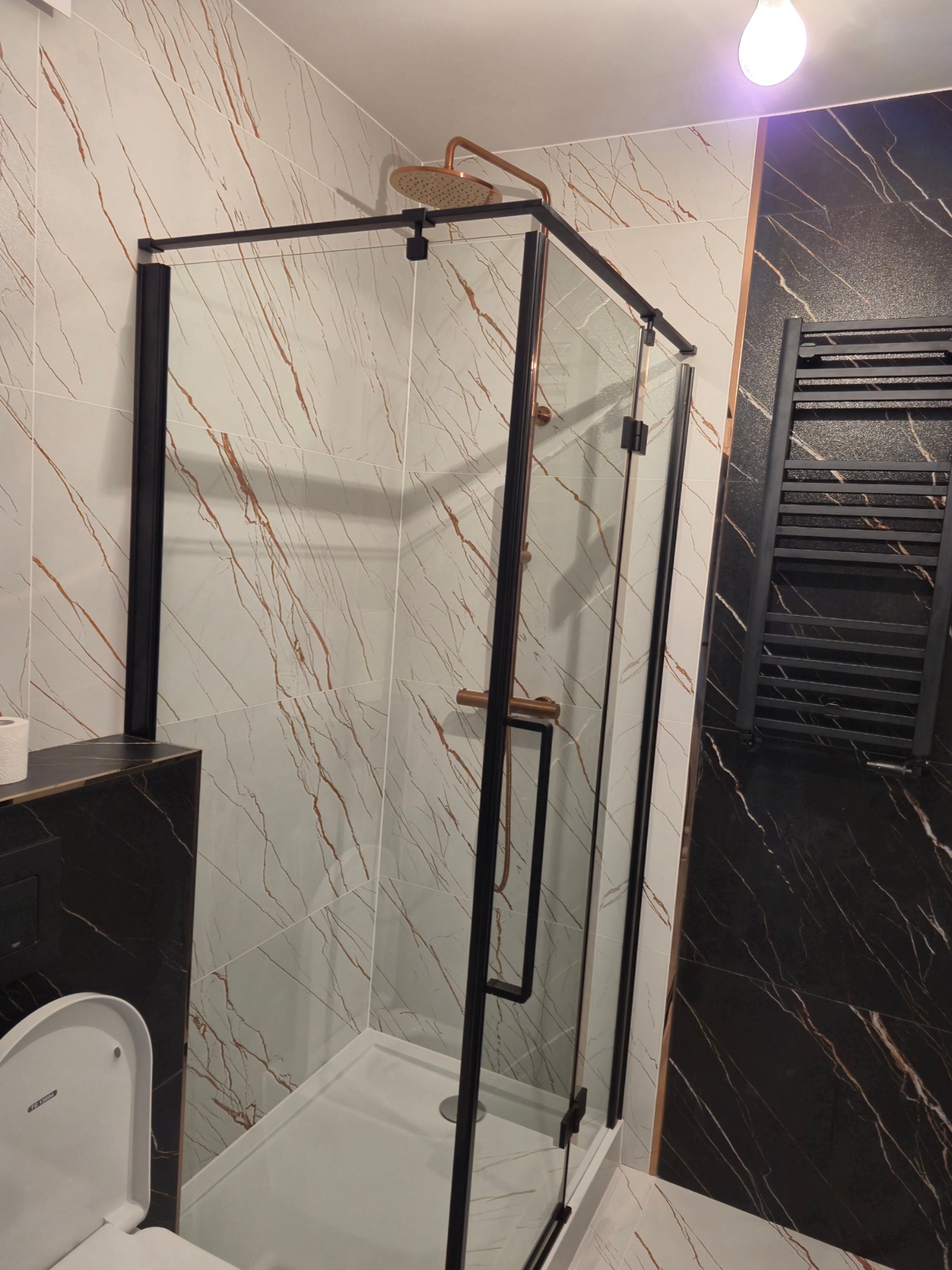 Nowoczesna kabina prysznicowa z czarną ramą i deszczownicą w kolorze miedzi, białe płytki z brązowymi żyłkami, czarny grzejnik drabinkowy na ścianie z czarnego marmuru, podświetlenie sufitowe.