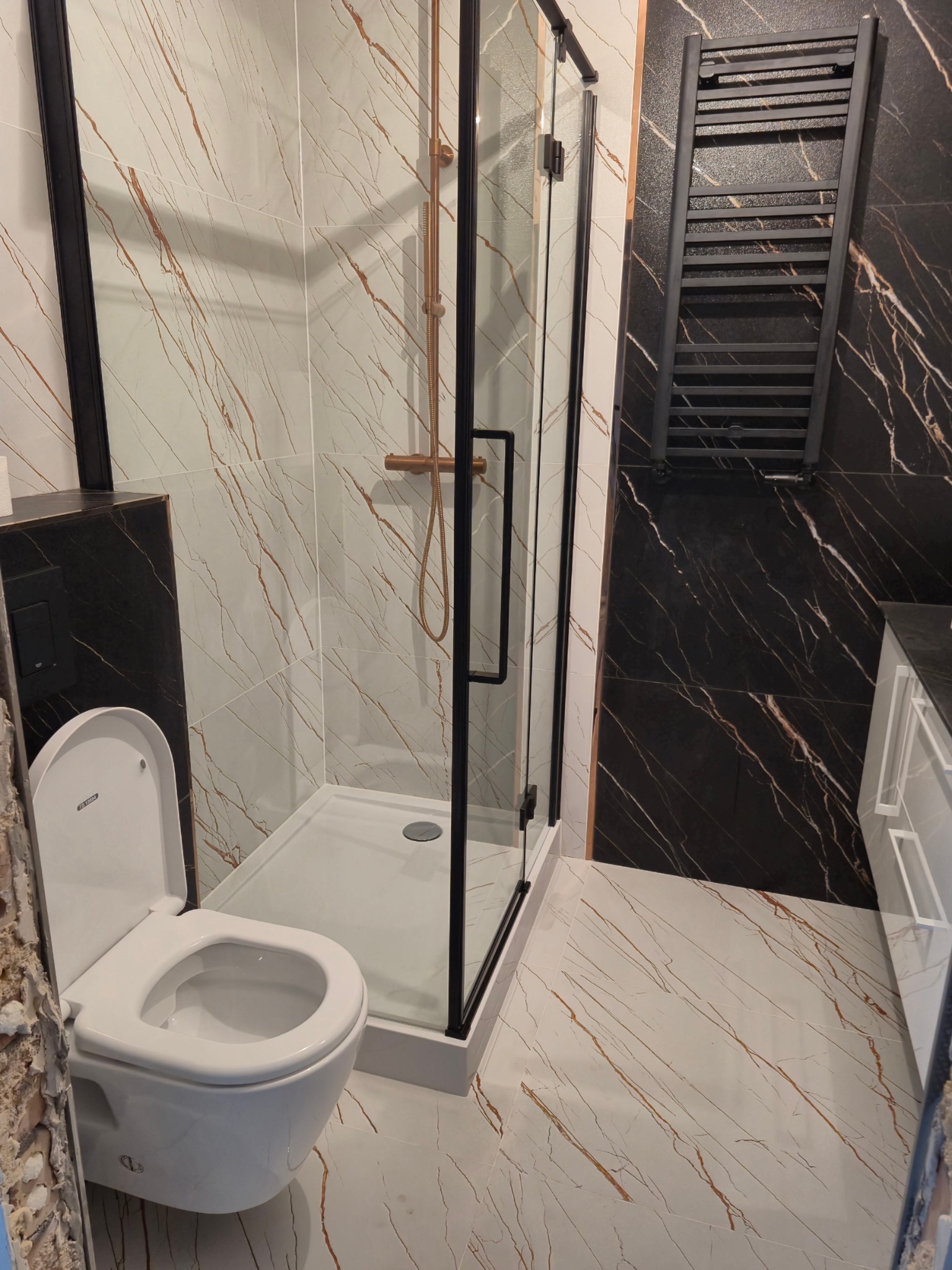 Nowoczesna łazienka w Gdańsku z kabiną prysznicową typu walk-in z czarnymi profilami, wiszącą toaletą i grzejnikiem drabinkowym na ścianie z czarnego marmuru.