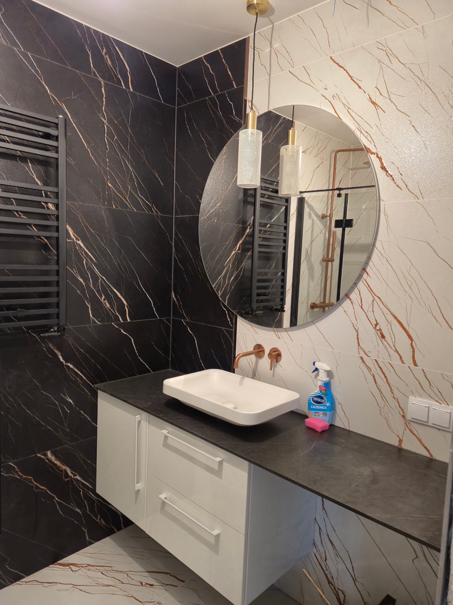 Nowoczesna łazienka z czarną i białą marmurkową płytką, okrągłym lustrem, wiszącą lampą z ażurowym kloszem, białą umywalką nablatową i baterią w kolorze miedzi oraz grzejnikiem drabinkowym.