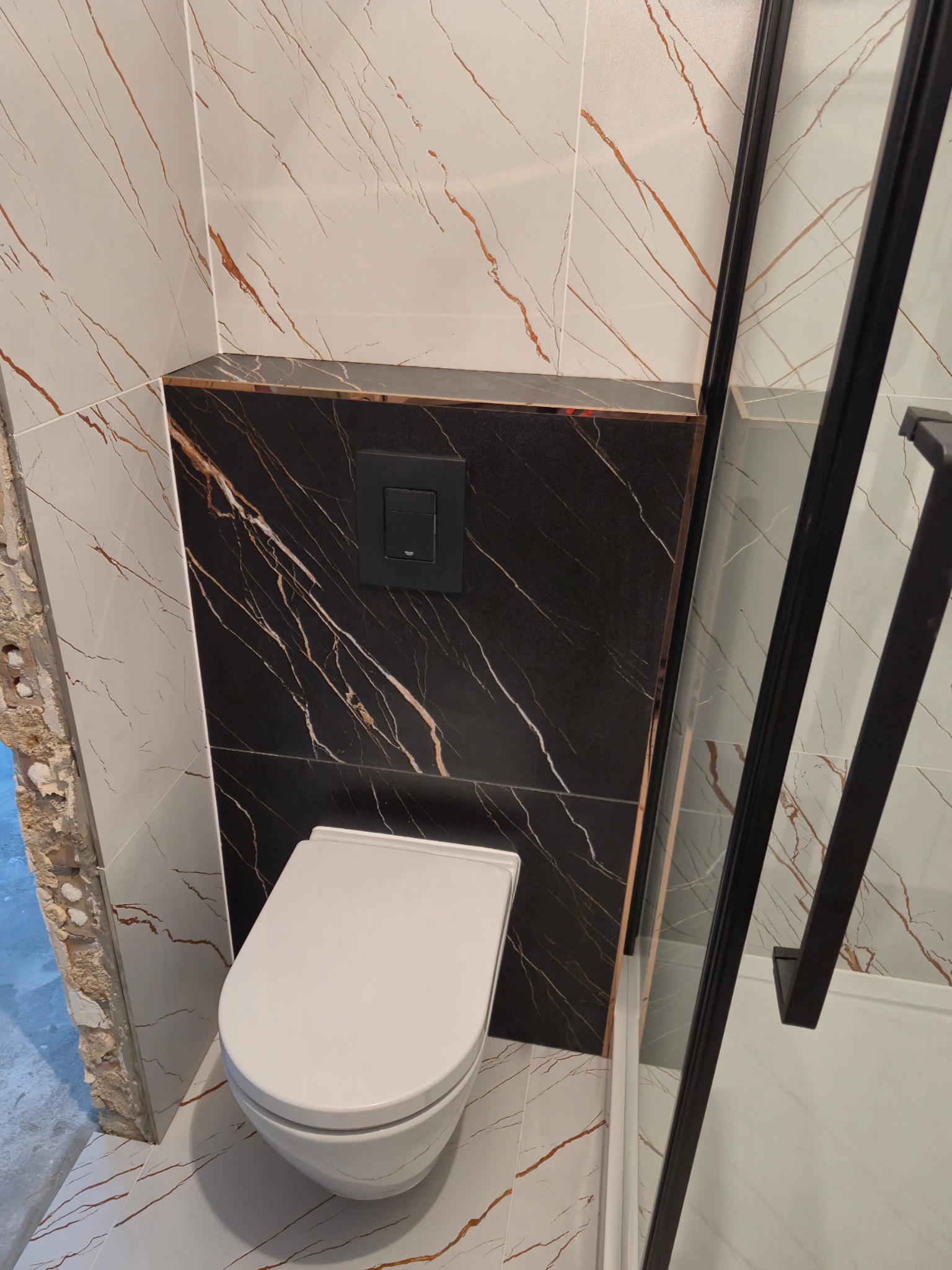 Podwieszana toaleta z czarnym panelem marmurowym, przy białych płytkach z brązowymi żyłkami i fragmentem kabiny prysznicowej z czarnymi profilami. Widoczne ślady po kuciu ściany.