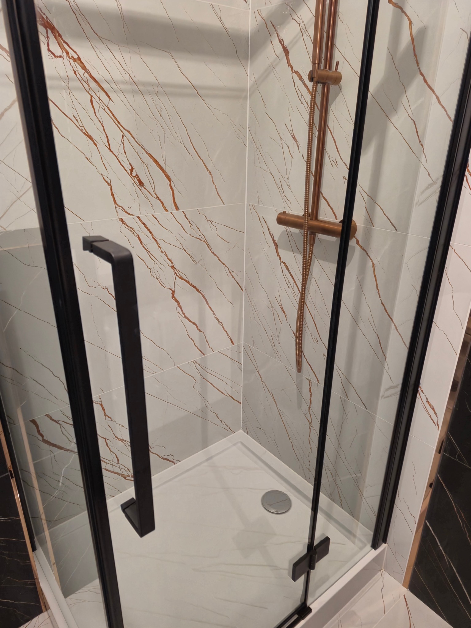 Nowoczesna kabina prysznicowa z czarną ramą i złotą armaturą na tle płytek imitujących marmur z brązowymi żyłkami.