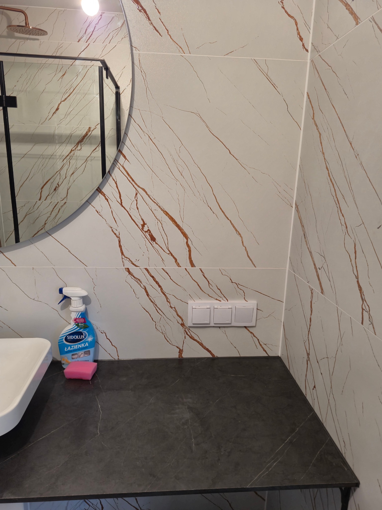 Umywalka z czarnym blatem, na którym leży różowa gąbka i stoi niebieski płyn do czyszczenia Sidolux Łazienka, w tle ściana wyłożona płytkami imitującymi marmur z brązowymi żyłami, nad blatem widać...