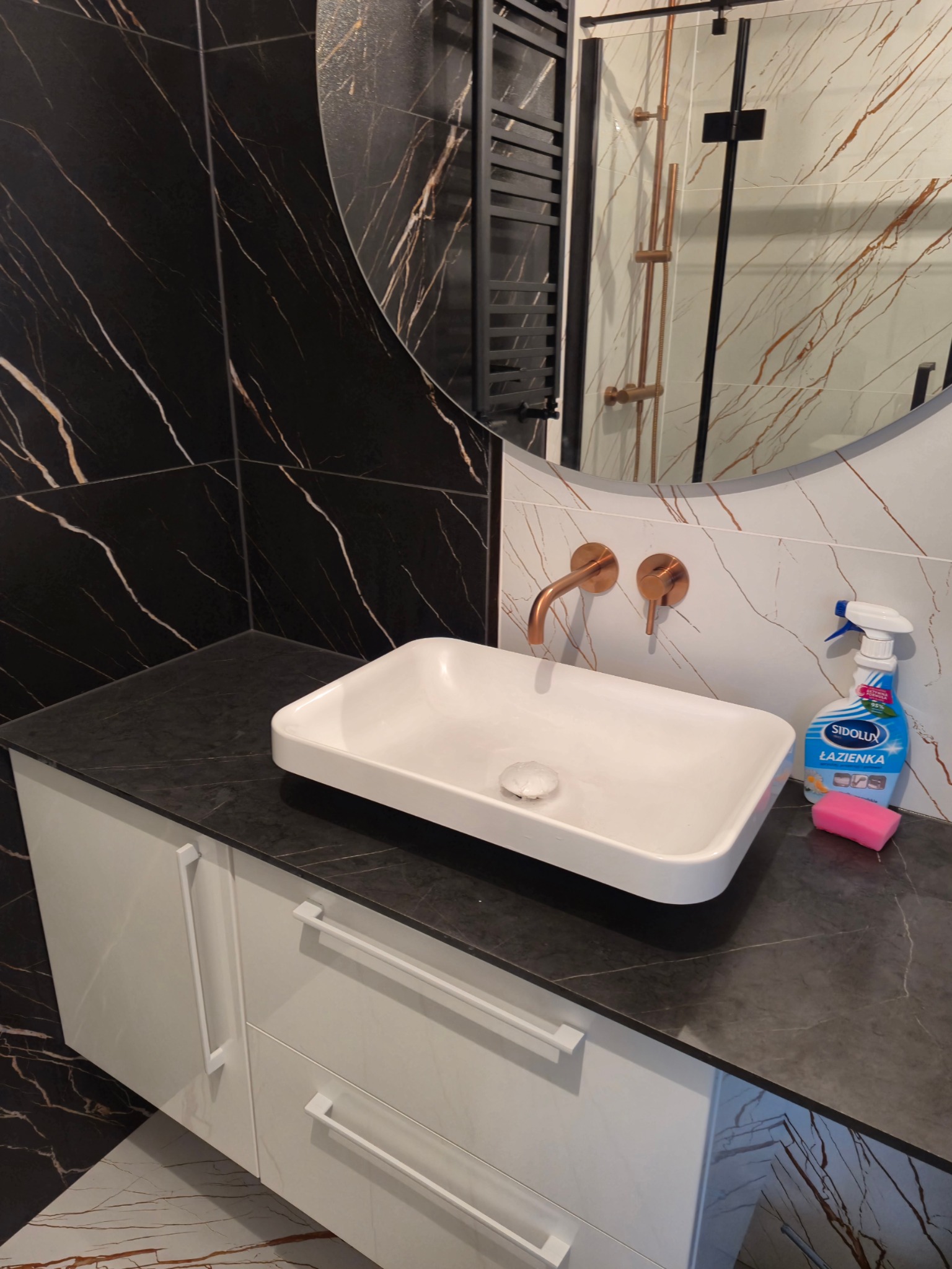 Nowoczesna łazienka z białą, prostokątną umywalką nablatową, miedzianą armaturą i czarnymi płytkami z marmurkowym wzorem. Widoczny fragment lustra, grzejnik drabinkowy i butelka płynu do czyszczenia.