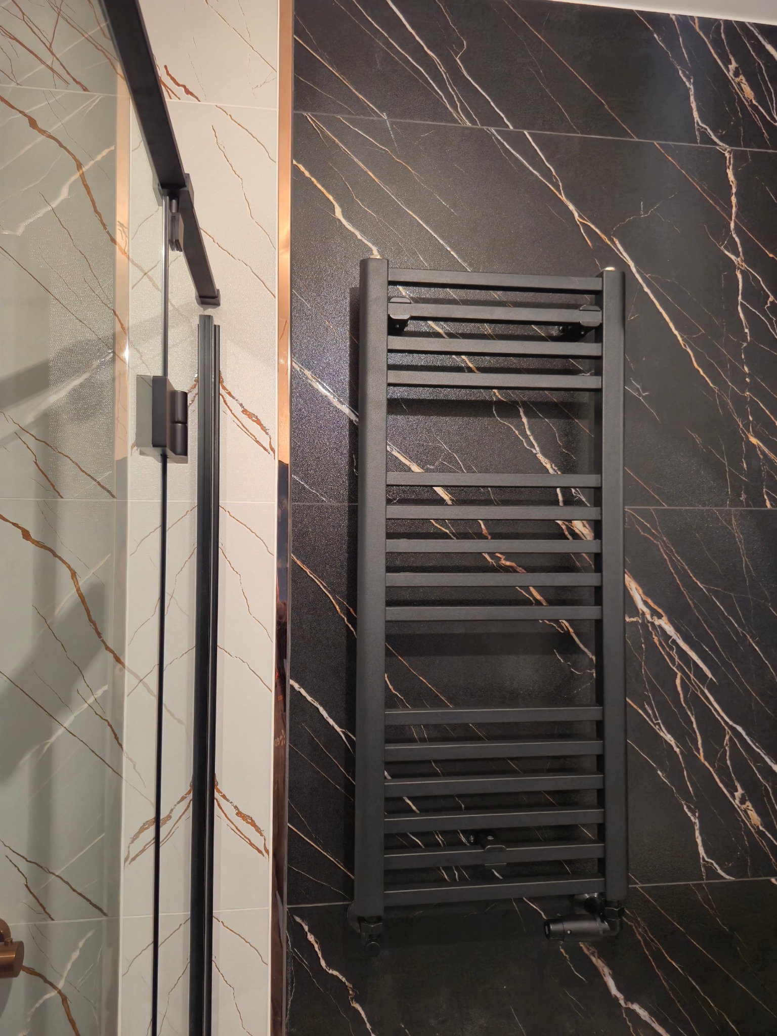 Czarny grzejnik drabinkowy na ścianie z czarnych płytek imitujących marmur, obok kabina prysznicowa z czarnymi profilami i białymi płytkami z marmurowym wzorem.