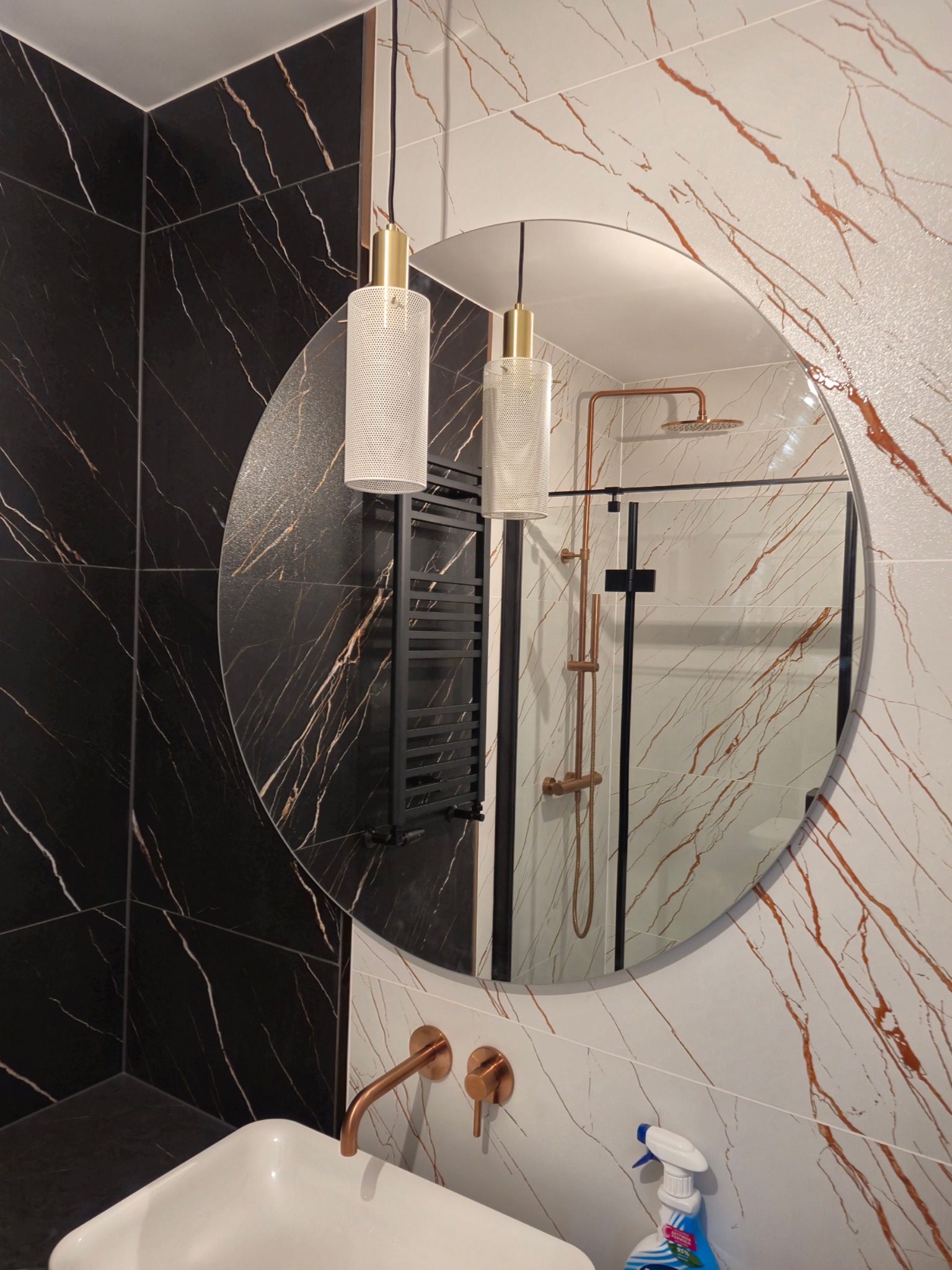 Nowoczesna łazienka z czarno-białymi płytkami imitującymi marmur, okrągłe lustro odbijające kabinę prysznicową z miedzianą armaturą, wiszące lampy z ażurowymi kloszami i biała umywalka z miedzianą...