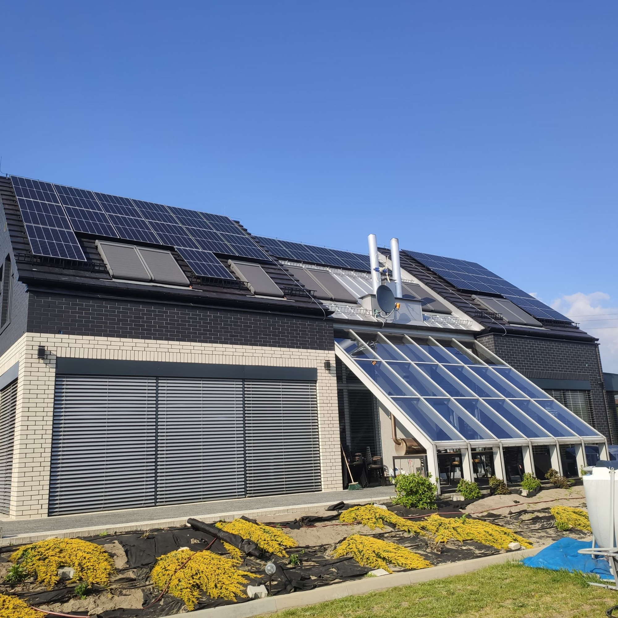 Nowoczesny dom z panelami słonecznymi na dachu, szklaną oranżerią i częściowo zagospodarowanym ogrodem z żółtymi krzewami.