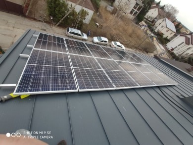 Panele słoneczne zamontowane na szarym dachu blaszanym z widokiem na okoliczne domy i zaparkowane samochody w pochmurny dzień.