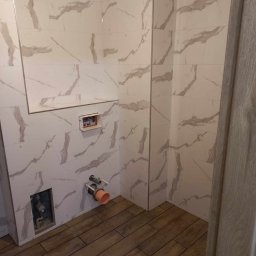 Artur Jurgielewicz - Wykończona łazienka z białymi płytkami imitującymi marmur na ścianach i drewnopodobną podłogą. Widoczne przyłącza wodno-kanalizacyjne i fragment ościeżnicy drzwi.