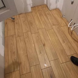 Artur Jurgielewicz - Ułożona podłoga z płytek imitujących drewno w małej łazience, widoczne szare ściany w trakcie wykończenia, fragment buta w rogu kadru.