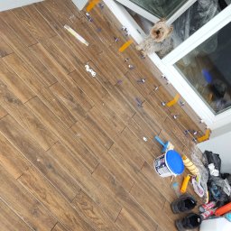 Artur Jurgielewicz - Układanie drewnopodobnych płytek podłogowych z użyciem poziomujących klipsów. Pies Yorkshire Terrier przy oknie obserwuje postęp prac.