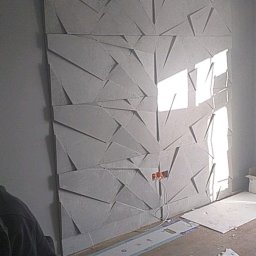 Artur Jurgielewicz - Montaż dekoracyjnej ściany 3D z paneli gipsowych. Widoczne narzędzia i fragment dłoni montera. Częściowo wykończona ściana z oświetleniem.