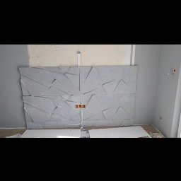 Artur Jurgielewicz - Montaż dekoracyjnych paneli ściennych 3D w szarym odcieniu. Widoczne gniazdka elektryczne i fragmenty płyt. Ściana pomalowana na szaro.