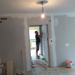 Artur Jurgielewicz - Mężczyzna montuje płytę gipsowo-kartonową w ościeżnicy podczas prac wykończeniowych, widoczne kable, puszka z gładzią i surowa żarówka na suficie.