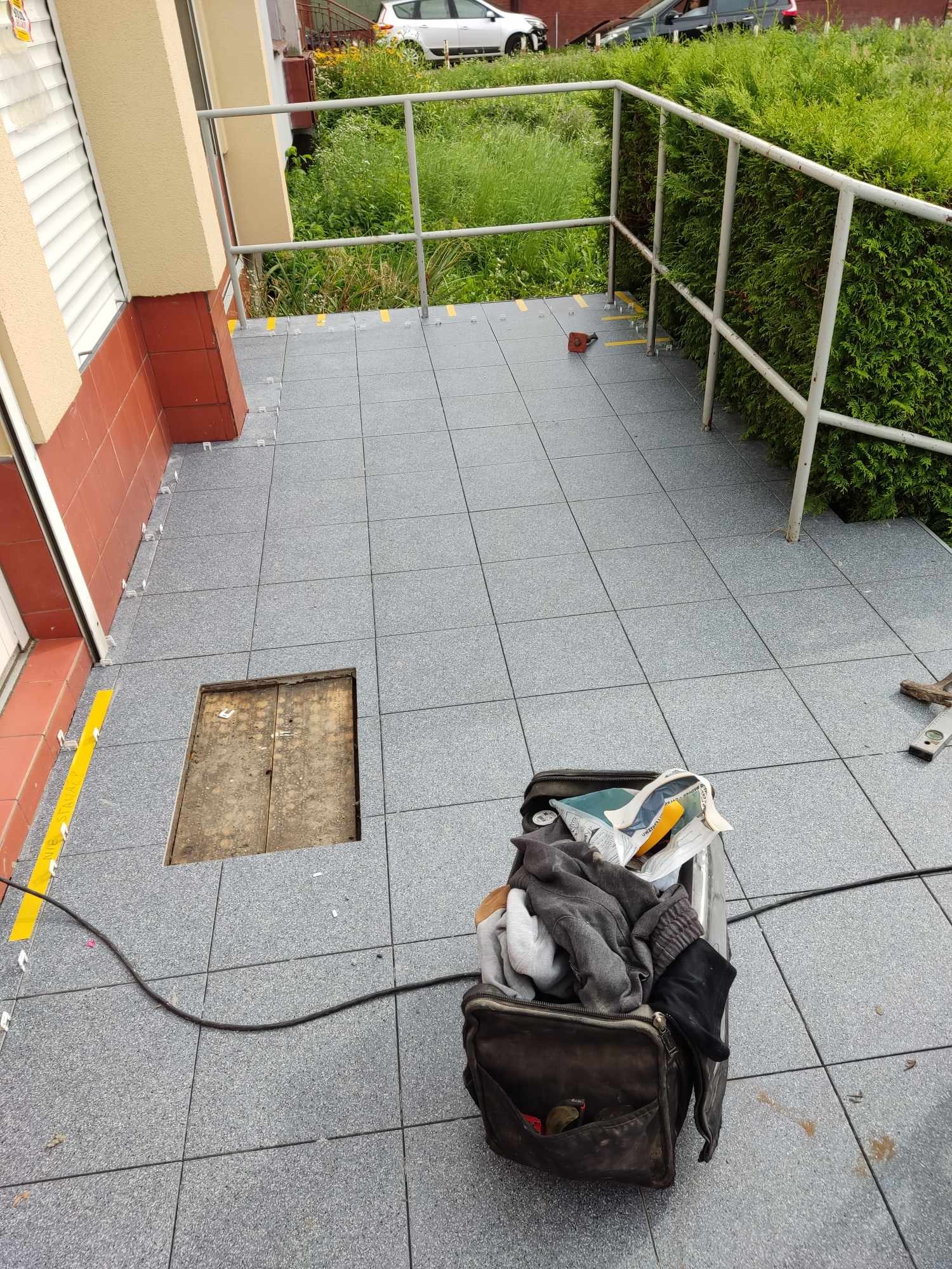 Montaż szarej terakoty na zewnątrz budynku, widoczne narzędzia pracy i fragment otwartego włazu. Fragment balustrady w tle.