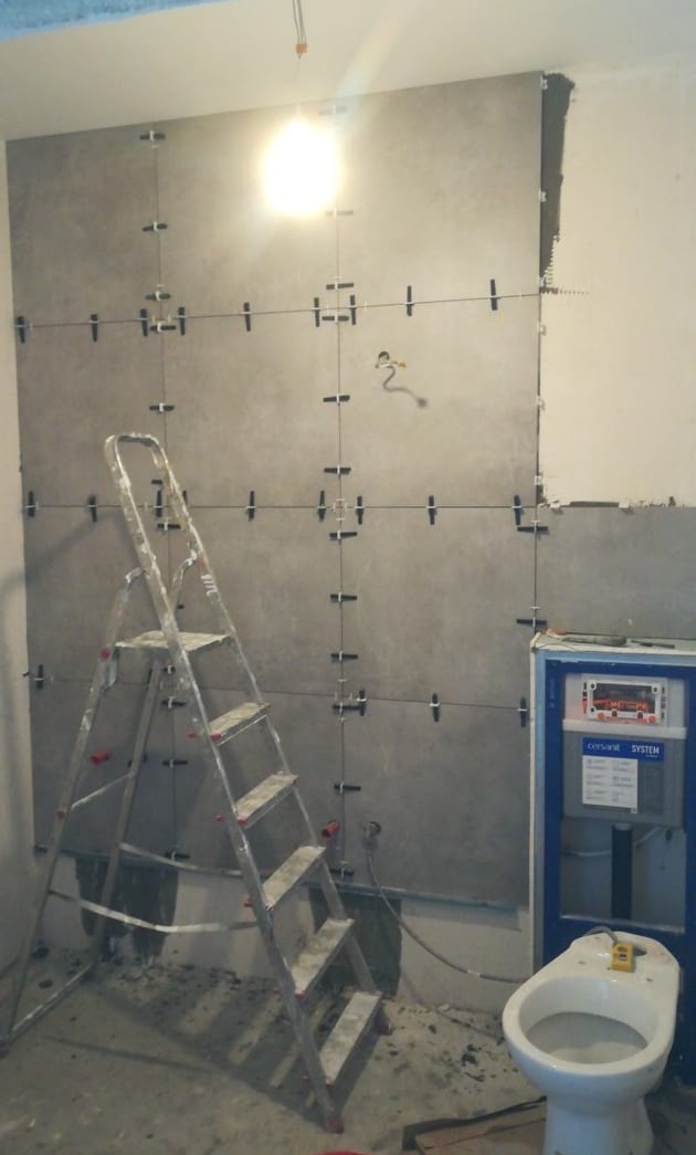 Ściana w trakcie układania płytek w łazience, widoczna drabina i toaleta. Użyte krzyżyki dystansowe. Szare płytki w dużym formacie.