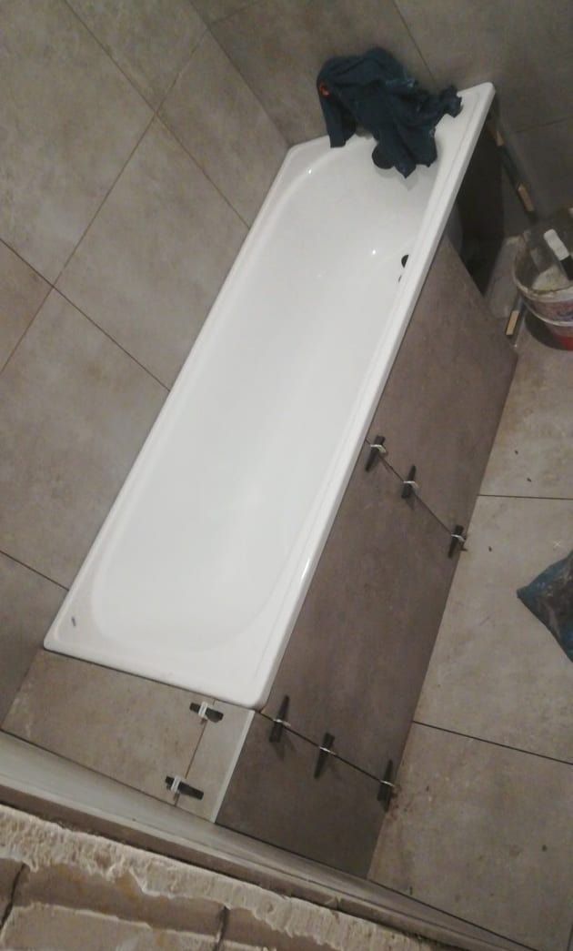 Biała wanna obudowana brązowymi płytkami, z widocznymi krzyżykami dystansowymi, w trakcie prac glazurniczych w łazience. Na wannie leżą ciemne ubrania.