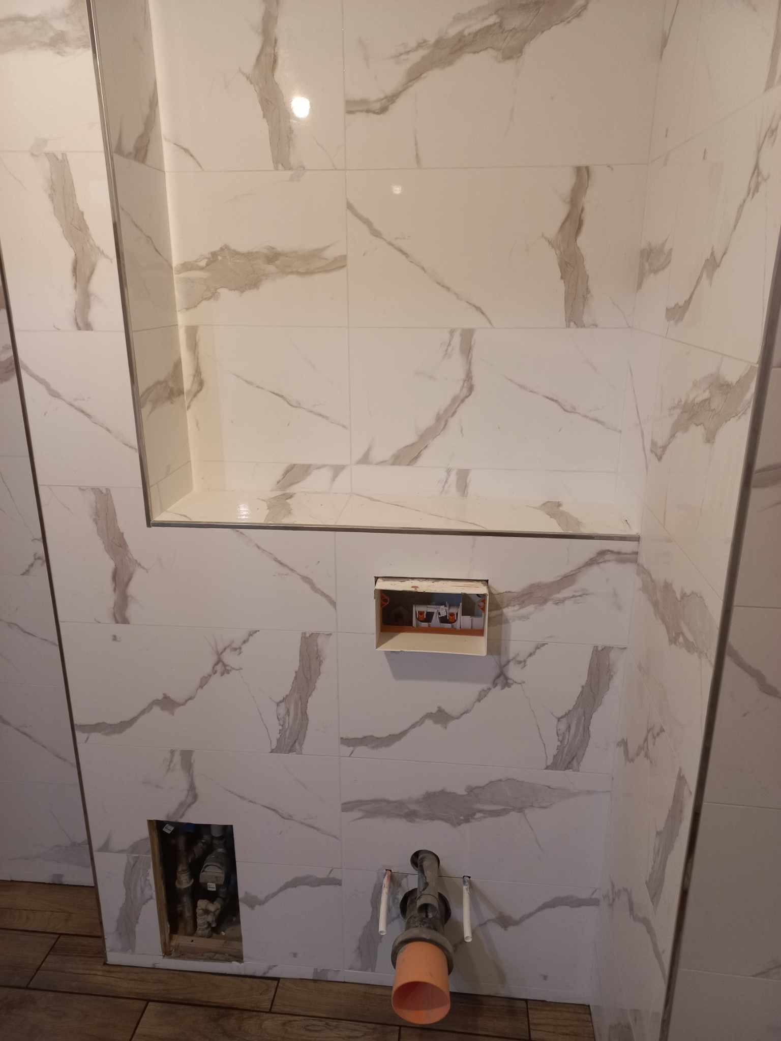 Wykończona wnęka i ściany z płytek imitujących marmur w łazience. Widoczne instalacje hydrauliczne i elektryczne przed montażem armatury.