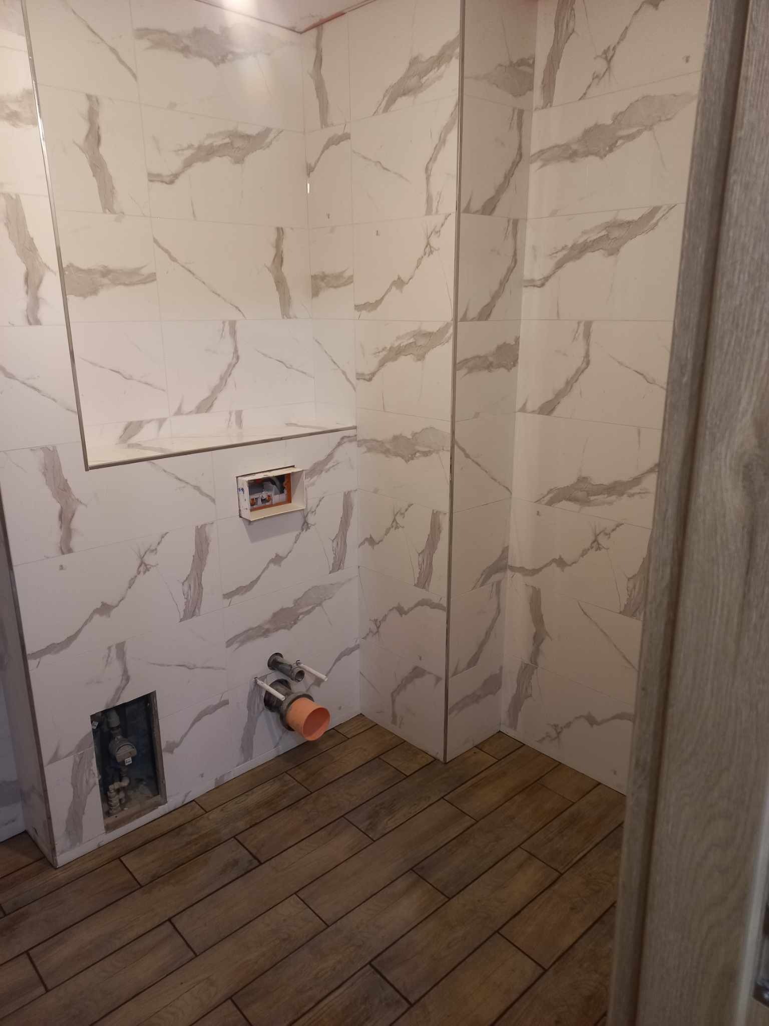 Wykończona łazienka z białymi płytkami imitującymi marmur na ścianach i drewnopodobną podłogą. Widoczne przyłącza wodno-kanalizacyjne i fragment ościeżnicy drzwi.