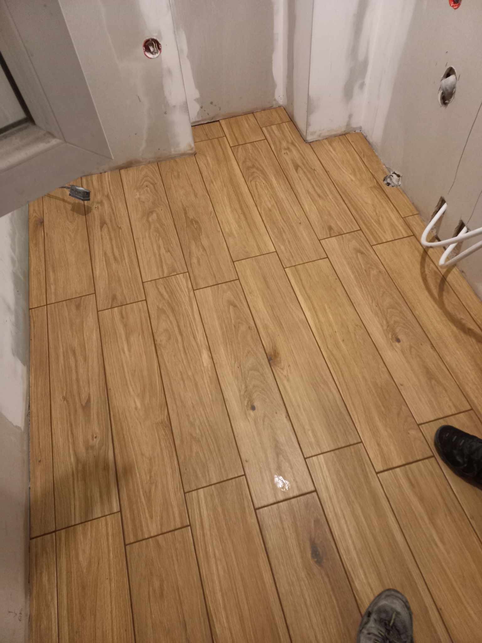 Ułożona podłoga z płytek imitujących drewno w małej łazience, widoczne szare ściany w trakcie wykończenia, fragment buta w rogu kadru.