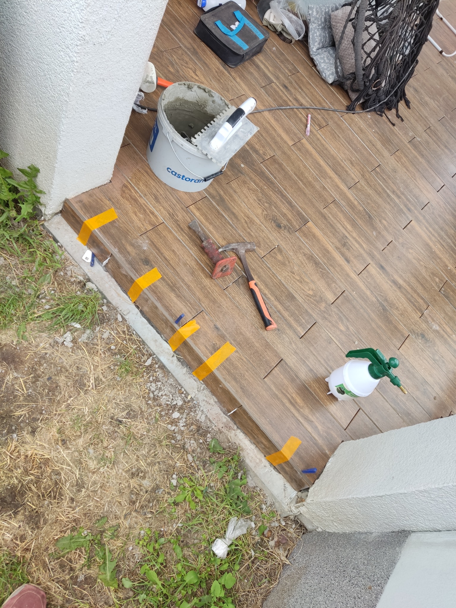 Układanie płytek imitujących drewno na zewnątrz, widoczne narzędzia glazurnicze, żółta taśma i poziomica. Prace w toku, widok z góry, naturalne światło.