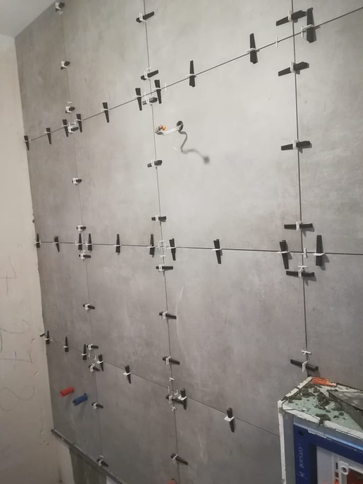 Montaż szarych płytek na ścianie z użyciem klipsów poziomujących. Widoczne przewody elektryczne i elementy instalacji wodnej. Prace glazurnicze w trakcie realizacji.