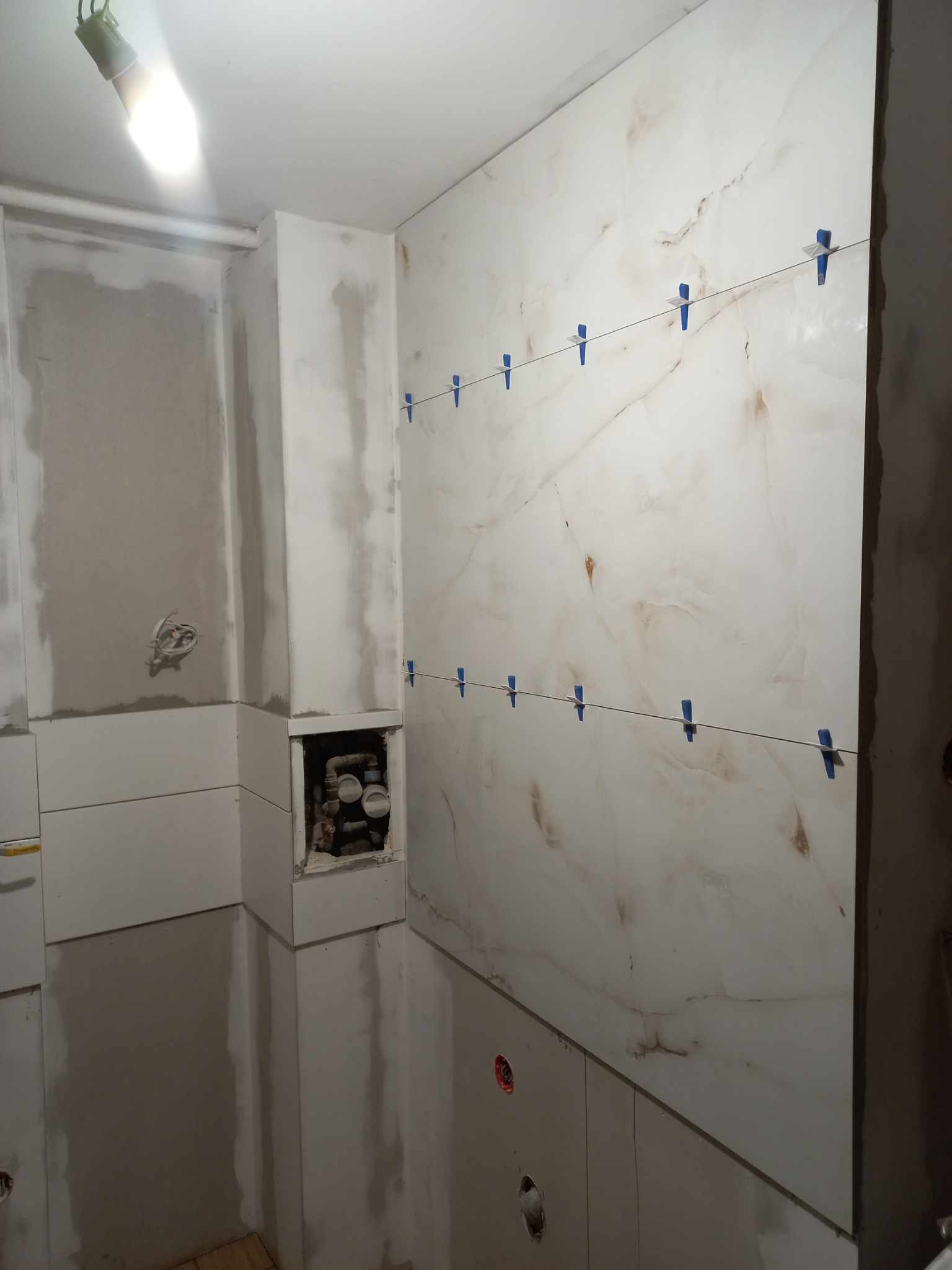 Układanie płytek marmuropodobnych w łazience, z użyciem niebieskich klipsów poziomujących. Widoczne instalacje. Oświetlenie sufitowe