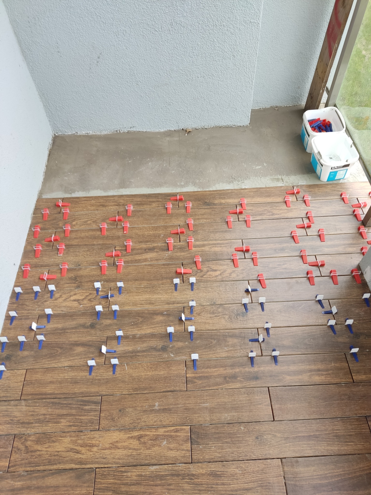 Układanie drewnopodobnych płytek na balkonie z użyciem poziomujących klipsów i klinów. Widoczne wiaderka z klejem i pozostałości zaprawy.