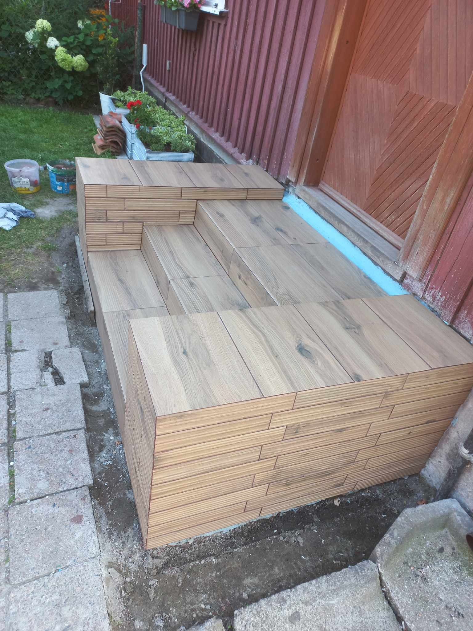 Ukończone schody wejściowe wyłożone płytkami imitującymi drewno, prowadzące do drewnianego domu z dekoracyjnym wzorem na drzwiach. Widoczne kwiaty i trawa w tle.