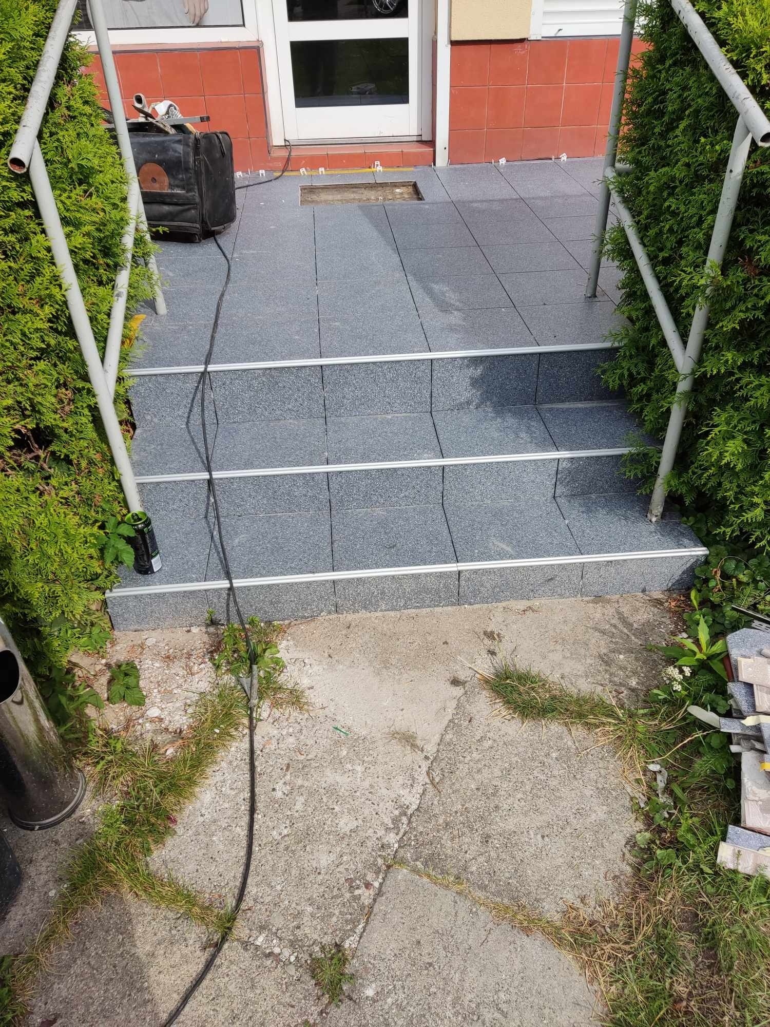 Schody wejściowe wyłożone szarymi płytkami z metalowymi listwami, widoczne narzędzia i niedokończone prace na górnym stopniu, otoczone zielenią.