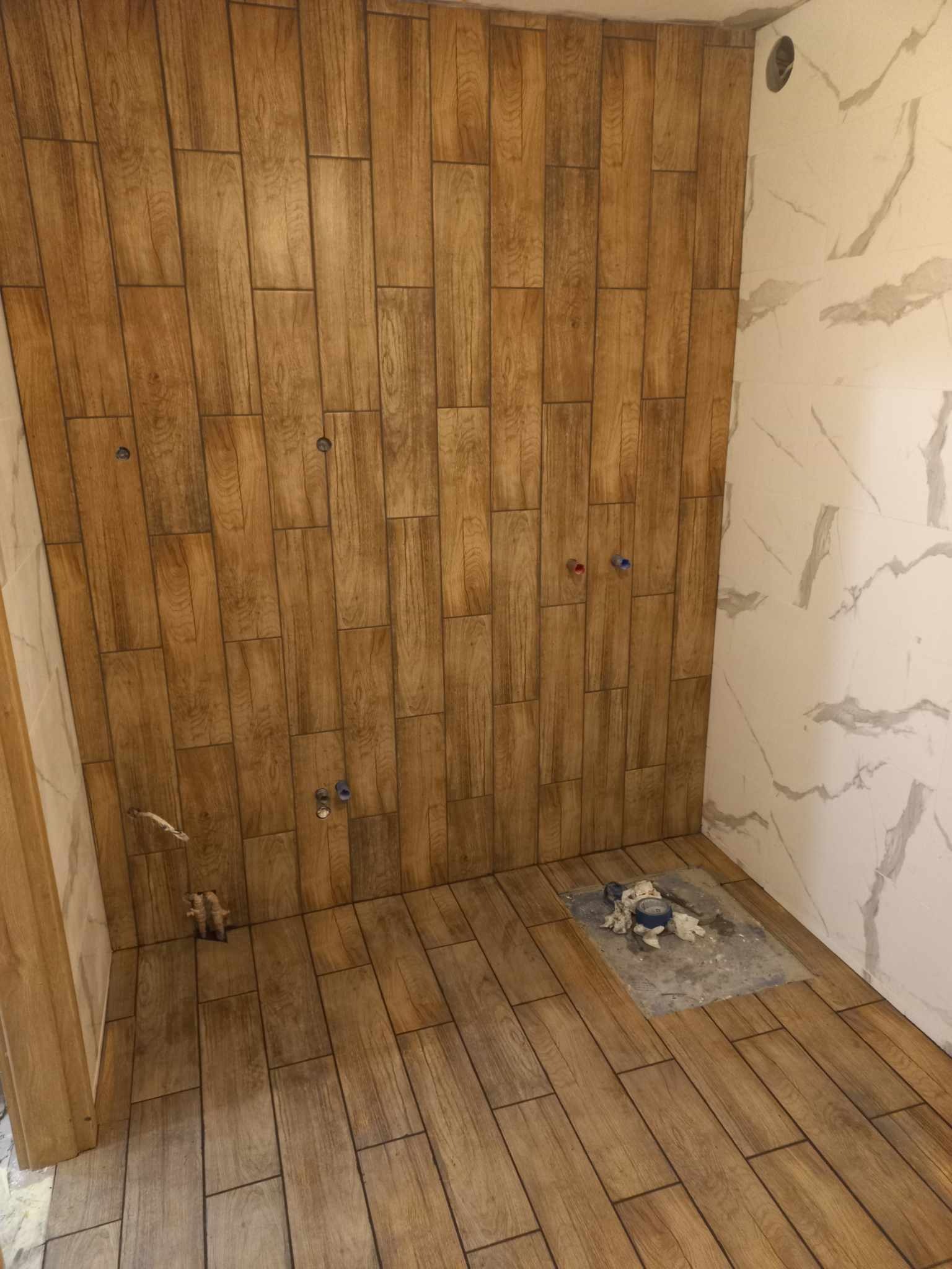 Wykończona ściana i podłoga w łazience z płytek imitujących drewno i marmur, widoczne przyłącza wodne, fragment instalacji pod umywalkę.