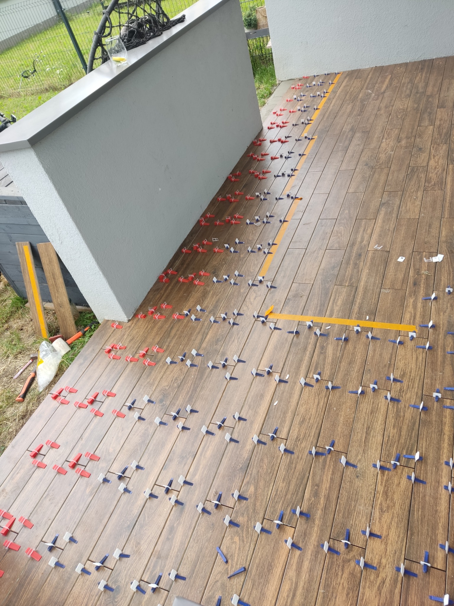 Układanie płytek imitujących drewno na zewnątrz, z użyciem klipsów poziomujących i taśmy. Widoczne narzędzia glazurnicze i fragment zieleni w tle.