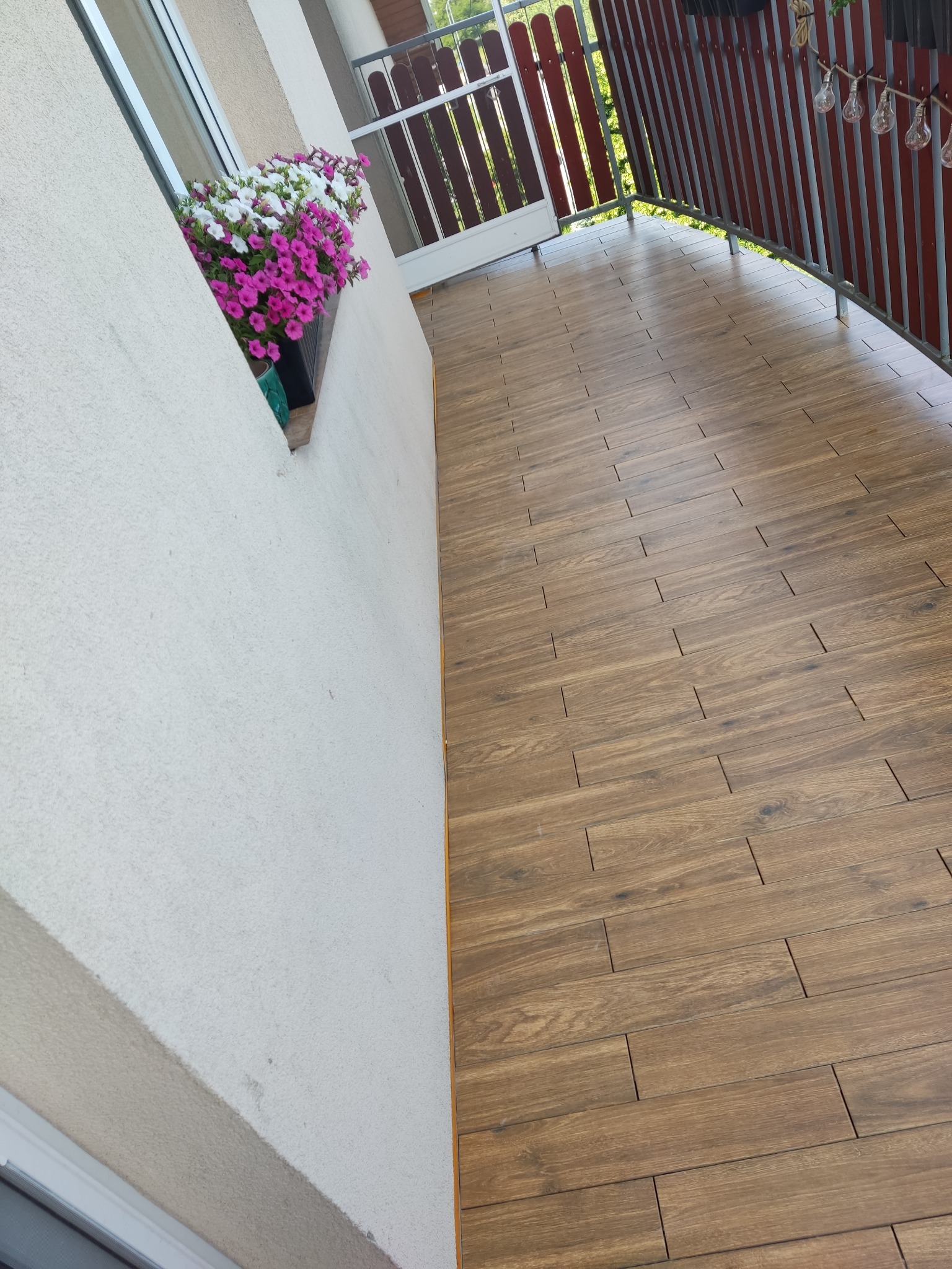 Balkon z drewnopodobną terakotą, kwiatami w oknie i dekoracyjnymi lampkami. Czerwona balustrada i widok na zieleń w tle.