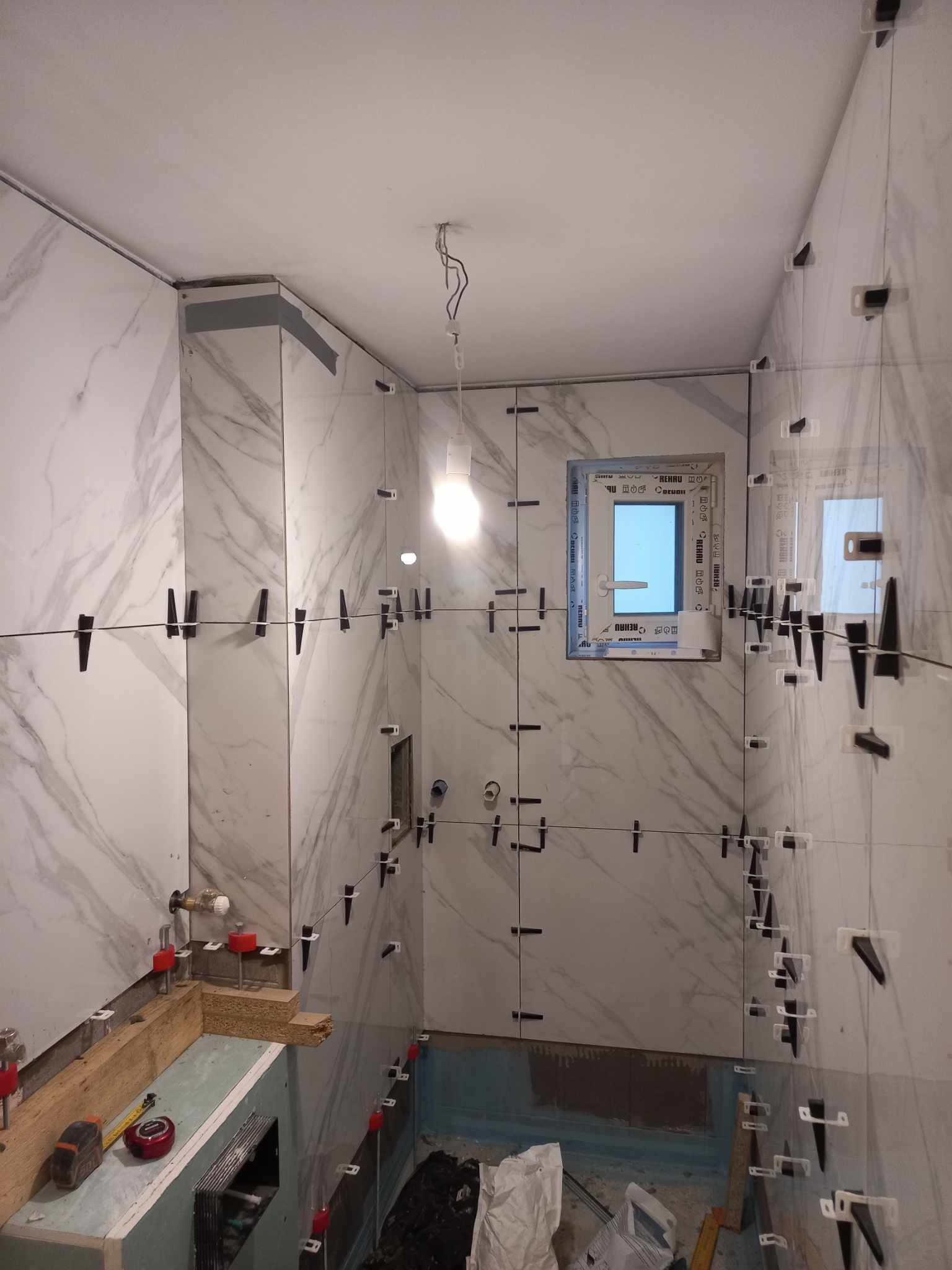 Układanie płytek imitujących marmur w łazience; widoczne krzyżyki dystansowe, okno i fragment przygotowanej zabudowy. Prace glazurnicze w toku.