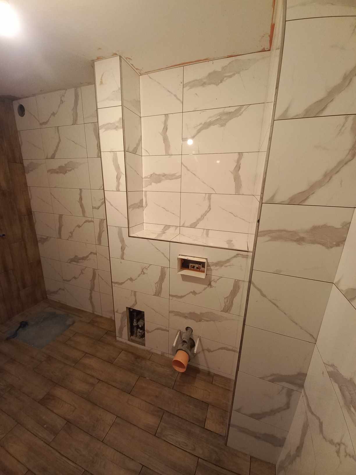 Wykończona łazienka z płytkami imitującymi marmur na ścianach i drewnopodobnymi na podłodze. Widoczne instalacje, wnęka w ścianie.