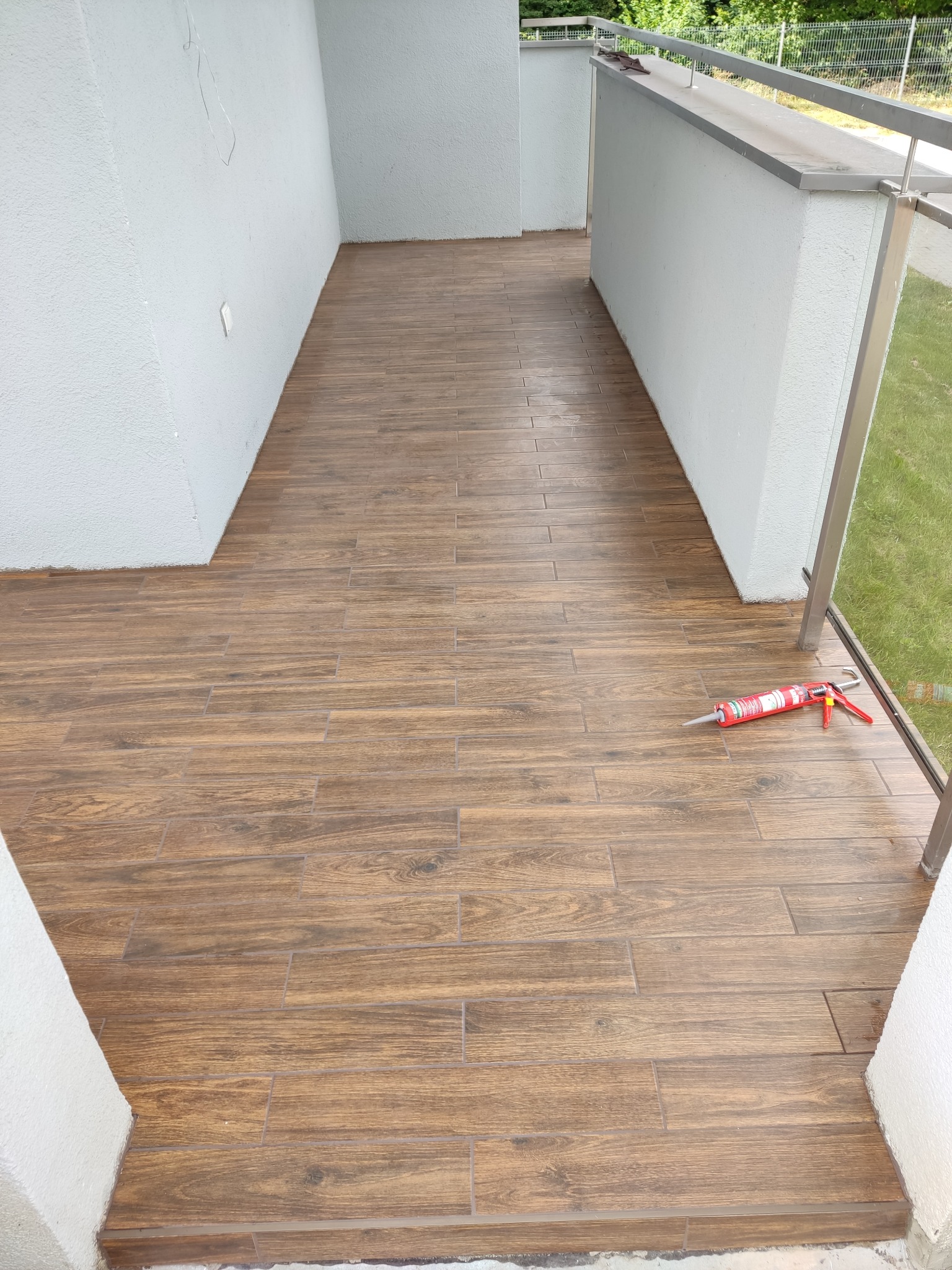 Balkon po remoncie z brązowymi płytkami imitującymi drewno. Widoczny aplikator do silikonu. Świeżo położona podłoga z płytek na balkonie.