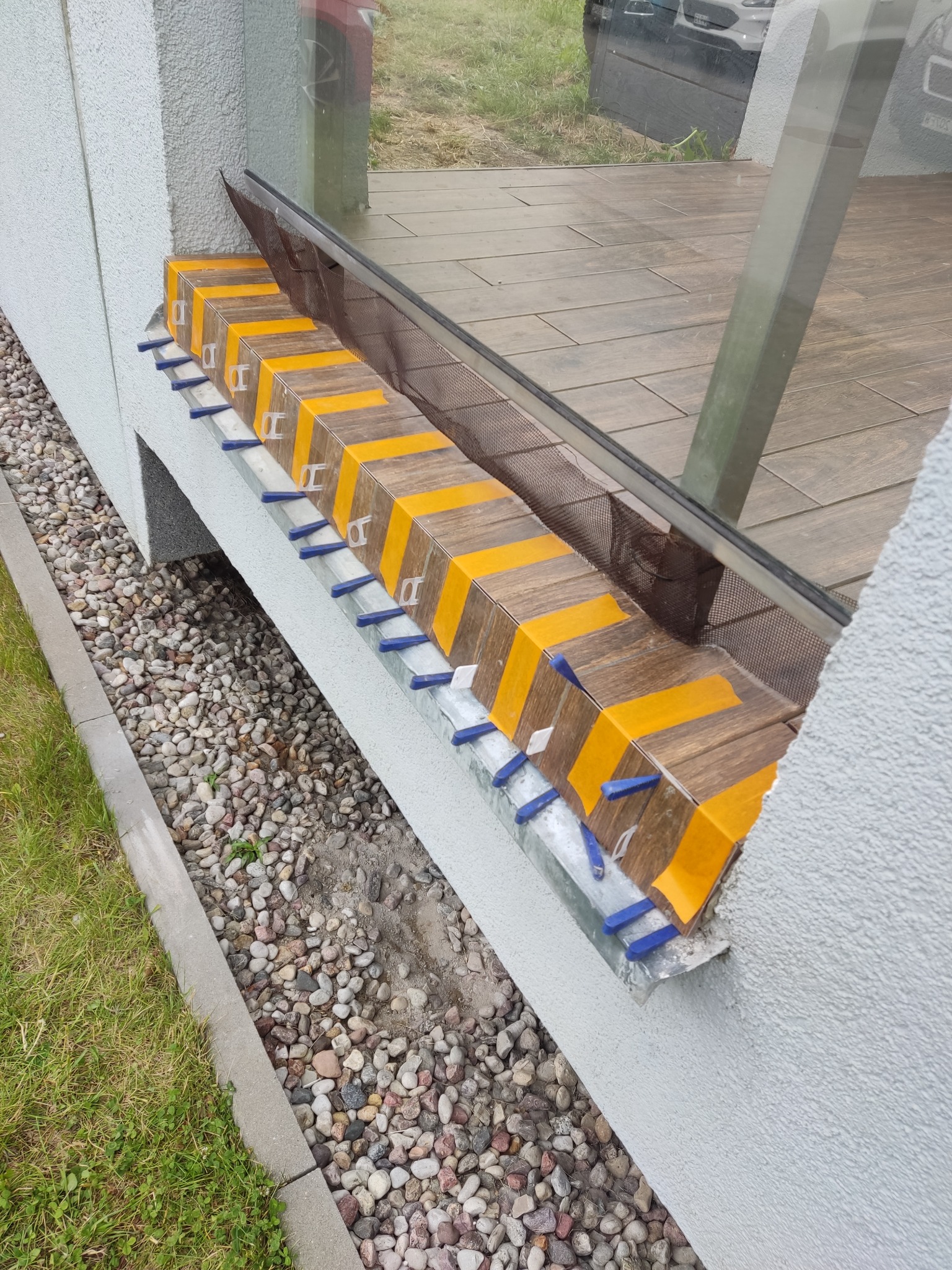 Montaż płytek imitujących drewno na zewnętrznym parapecie okiennym. Użyto niebieskich klinów dystansowych i żółtej taśmy. Widoczne kamienie dekoracyjne i trawa.