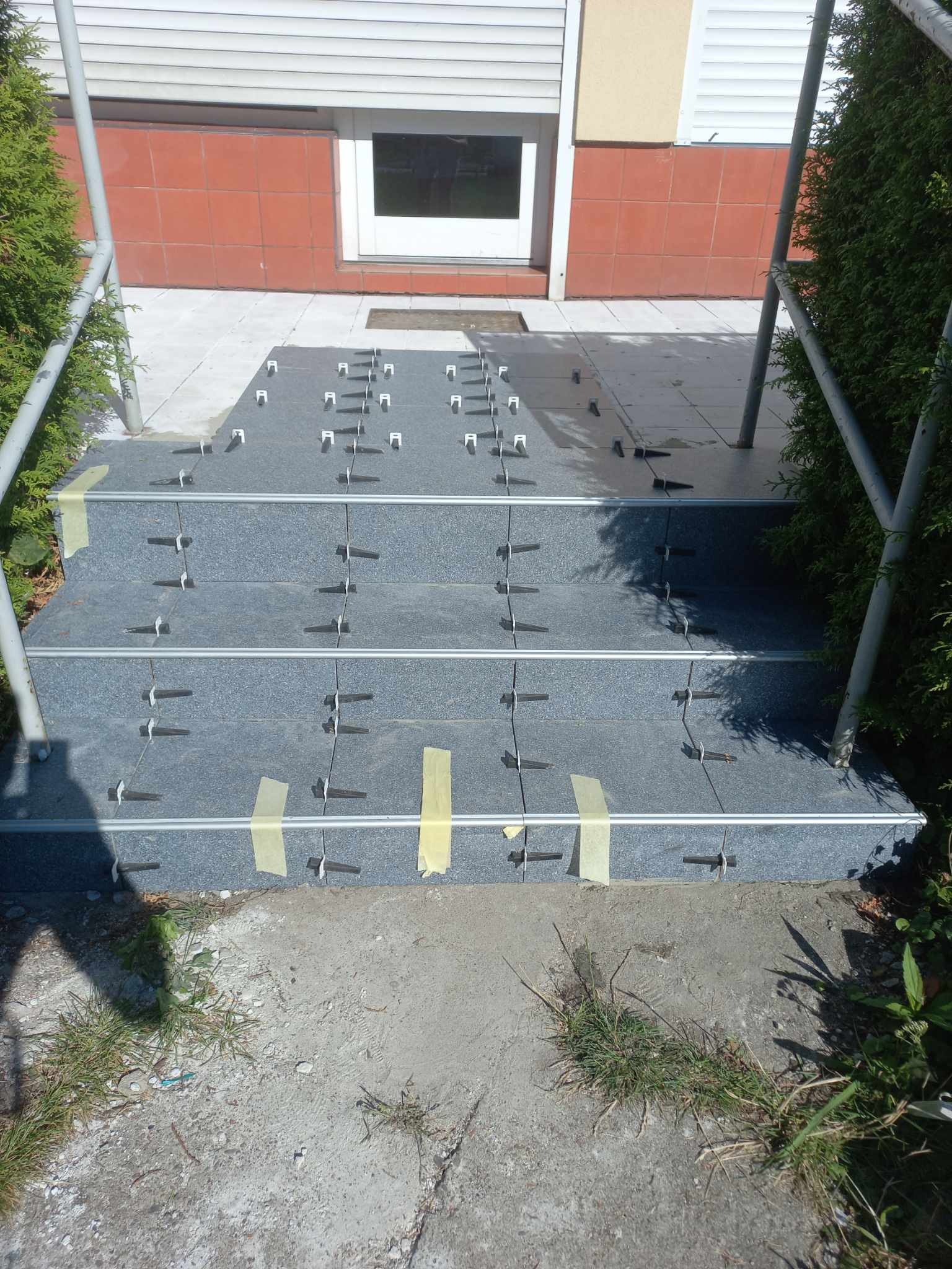 Kładzenie szarych płytek na schodach wejściowych, użycie klipsów poziomujących i taśmy. Widoczne balustrady i roślinność. Prace glazurnicze w toku.