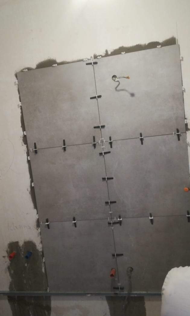 Układanie dużych, szarych płytek z krzyżykami dystansowymi na ścianie w łazience, widoczne przewody elektryczne i fragment instalacji wodnej.