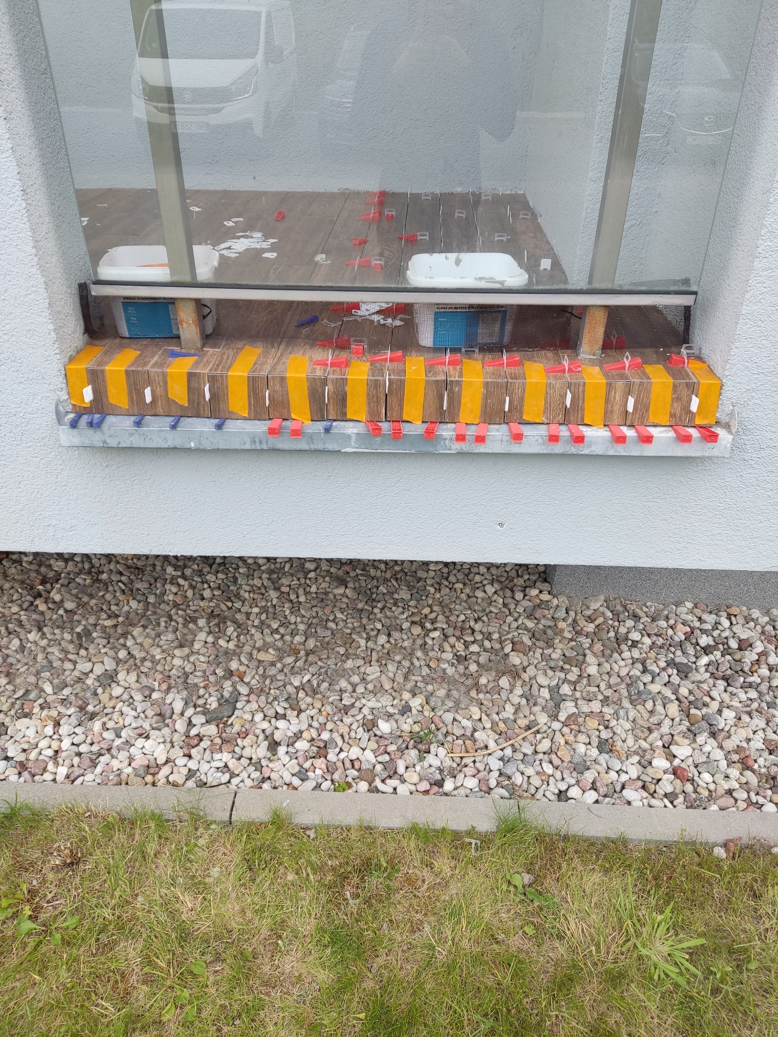 Układanie płytek na parapecie zewnętrznym z użyciem poziomowania i taśmy. Widoczne narzędzia glazurnicze i fragment trawnika.