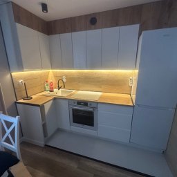 ZAKŁAD IZOLACJI I USŁUG BUDOWLANYCH Artur Marach - Nowoczesna, jasna kuchnia z białymi szafkami i drewnianym blatem. Widoczny fragment krzesła. Oświetlenie LED pod szafkami górnymi. Minimalistyczny design.