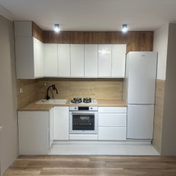 ZAKŁAD IZOLACJI I USŁUG BUDOWLANYCH Artur Marach - Nowoczesna kuchnia z białymi szafkami, drewnianymi akcentami i sprzętem AGD. Uwagę przykuwa drewniana ściana nad blatem i podświetlenie LED. Podłoga wyłożona panelami imitującymi drewno.