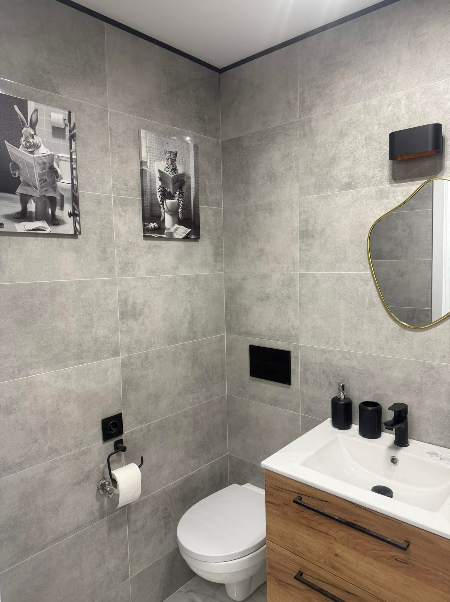Nowoczesna łazienka z szarymi płytkami, drewnianą szafką pod umywalką i lustrem w złotej ramie. Na ścianie wiszą czarno-białe grafiki z motywami zwierzęcymi.