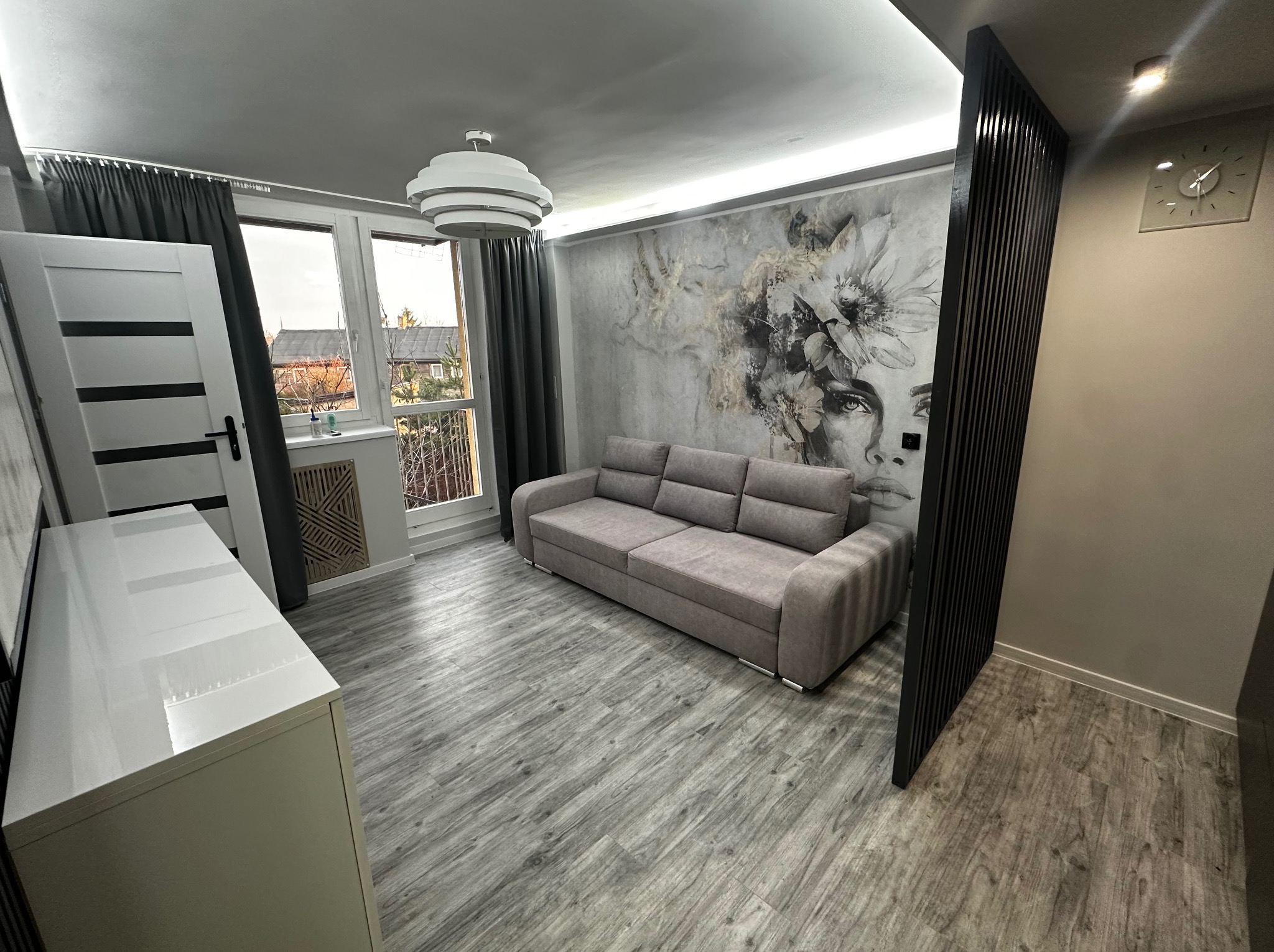 Jasne wnętrze pokoju z szarą sofą, panelami dekoracyjnymi i fototapetą z motywem twarzy. Widoczne okno z zasłonami i element białej szafki.