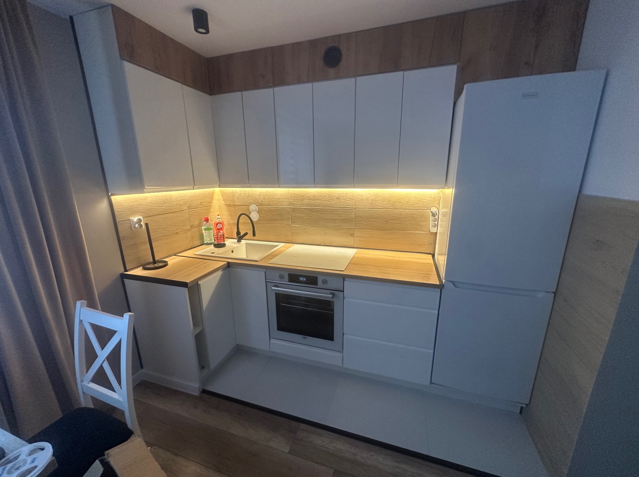 Nowoczesna, jasna kuchnia z białymi szafkami i drewnianym blatem. Widoczny fragment krzesła. Oświetlenie LED pod szafkami górnymi. Minimalistyczny design.