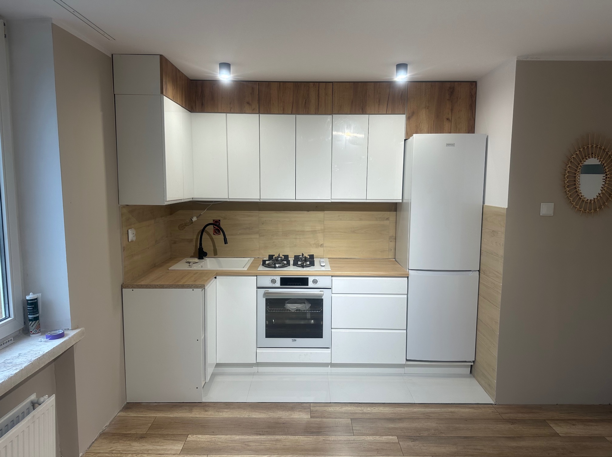 Nowoczesna kuchnia z białymi szafkami, drewnianymi akcentami i sprzętem AGD. Uwagę przykuwa drewniana ściana nad blatem i podświetlenie LED. Podłoga wyłożona panelami imitującymi drewno.