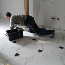 BRYGADA102.PL SPÓŁKA Z OGRANICZONĄ ODPOWIEDZIALNOŚCIĄ - Mężczyzna w roboczym ubraniu układa białe płytki podłogowe z czarnymi wstawkami w pomieszczeniu z surowymi, drewnianymi elementami konstrukcyjnymi i niezamaskowanymi instalacjami wodnymi na ścianie.