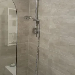 Nowoczesna łazienka z beżowymi płytkami imitującymi drewno, kabiną prysznicową z hartowanego szkła i chromowaną armaturą, w tym baterią wannową i zestawem prysznicowym z deszczownicą.