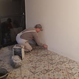 BRYGADA102.PL SPÓŁKA Z OGRANICZONĄ ODPOWIEDZIALNOŚCIĄ - Mężczyzna w kombinezonie i czapce, na kolanach, wykańcza fugi przy podłodze z wzorzystych płytek ceramicznych, przy ścianie, widoczny grzejnik elektryczny i wiadro z narzędziami.