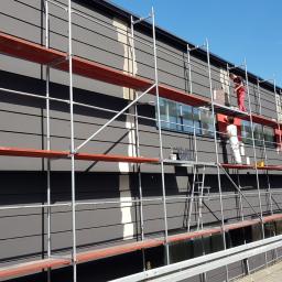 BRYGADA102.PL SPÓŁKA Z OGRANICZONĄ ODPOWIEDZIALNOŚCIĄ - Malowanie elewacji budynku z rusztowania: dwóch malarzy w kombinezonach, jeden w czerwonym, drugi w białym, aplikują farbę na szarą elewację z poziomymi panelami, widoczne okna i fragment chodnika.